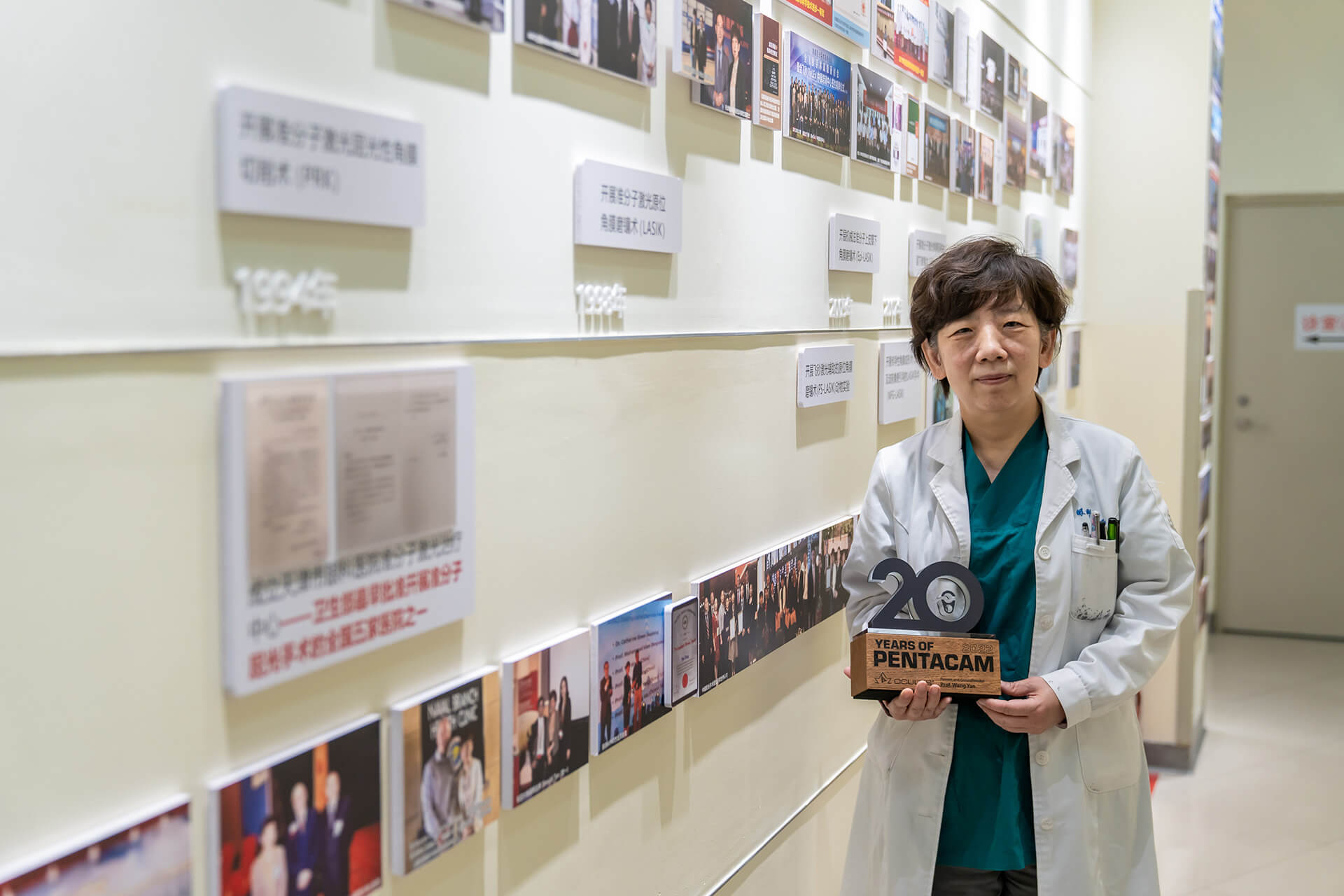 Prof. Wang Yan hält ihre Pentacam®-Trophäe zum 20-jährigen Jubiläum, und die Rückwand zeigt die Entwicklung ihres Labors für refraktive Chirurgie seit mehr als 20 Jahren